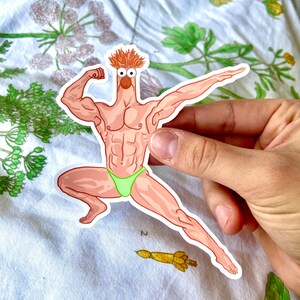 May include: A sticker featuring a cartoon character with a muscular physique, orange hair, and a green swimsuit. The character is flexing its arm and striking a pose. The sticker is held by a hand.