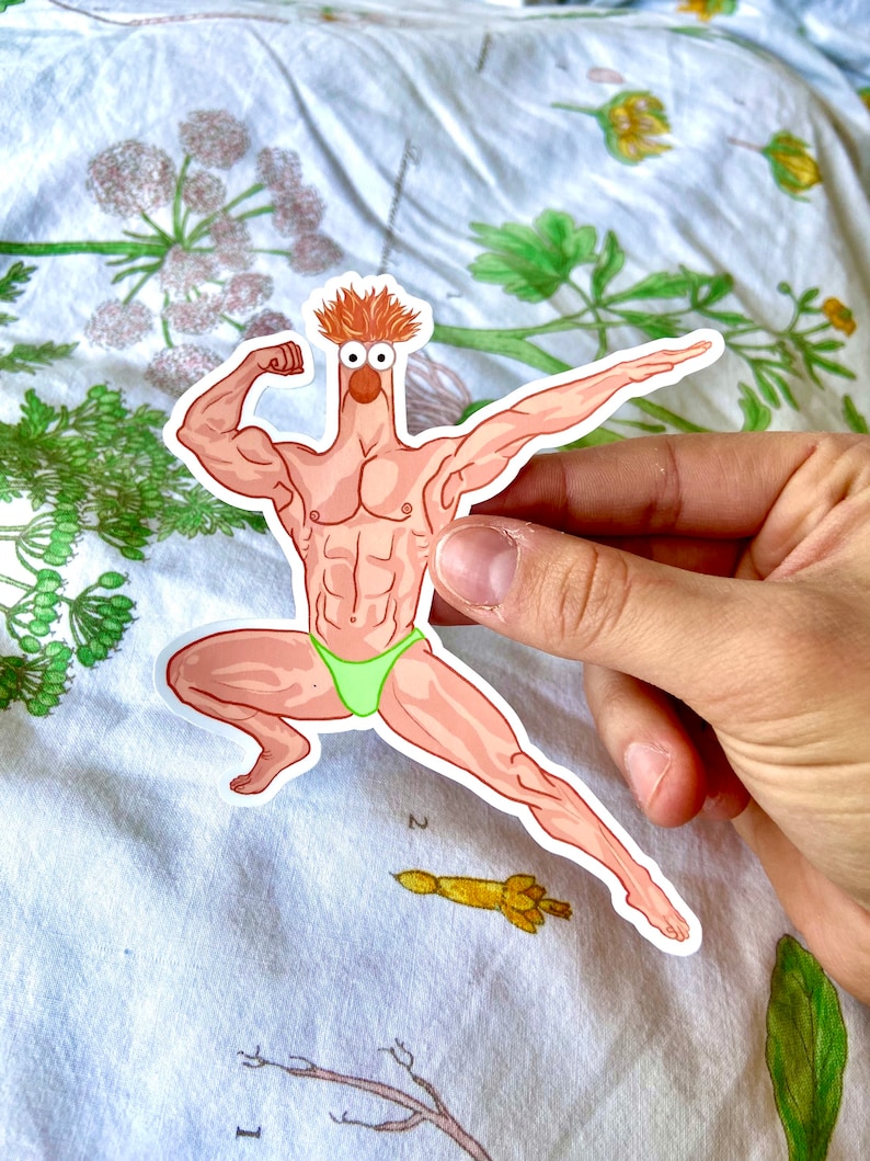 May include: A sticker featuring a cartoon character with a muscular physique, orange hair, and a green swimsuit. The character is flexing its arm and striking a pose. The sticker is held by a hand.