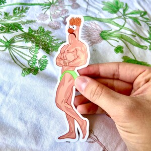 May include: A sticker featuring a cartoon character with a muscular physique, wearing green briefs. The character has orange hair, a red nose, and is standing with arms crossed. The sticker is held by a hand against a floral background.