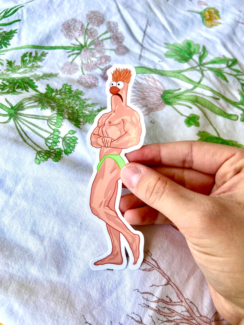 May include: A sticker featuring a cartoon character with a muscular physique, wearing green briefs. The character has orange hair, a red nose, and is standing with arms crossed. The sticker is held by a hand against a floral background.