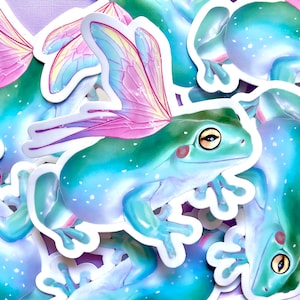 May include: A set of colorful frog stickers with iridescent wings. The frogs are green, blue, and purple with white spots. The wings are pink, blue, and yellow.