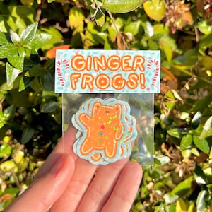 May include: A clear plastic bag with a sticker that says "Ginger Frogs!" and features a gingerbread frog with colorful sprinkles. The sticker is on a blue background with white candy cane designs.
