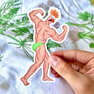 May include: A sticker featuring a cartoon character with a muscular physique, orange hair, and a red nose. The character is wearing green briefs and flexing its arms. The sticker is held by a hand against a floral background.
