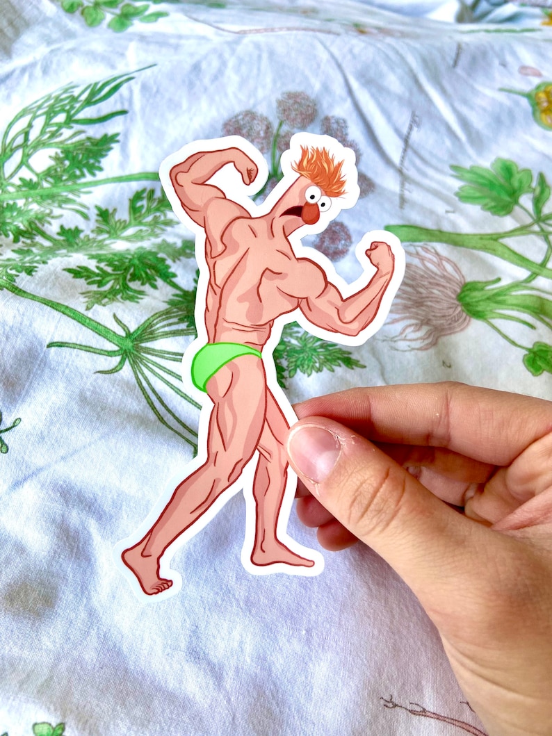 May include: A sticker featuring a cartoon character with a muscular physique, orange hair, and a red nose. The character is wearing green briefs and flexing its arms. The sticker is held by a hand against a floral background.