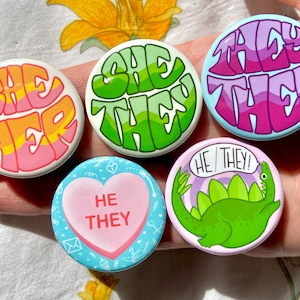 Puede incluir: Cinco botones coloridos con diferentes diseños. Los botones muestran las frases "She Her", "She They", "They Them", "He They!" y un corazón rosa con el texto "He They".
