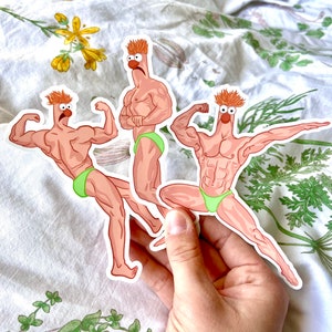 May include: Three stickers featuring a cartoon character with a muscular physique, orange hair, and a red nose. The character is wearing green briefs and striking bodybuilding poses. The stickers are held in a hand against a floral background.
