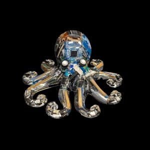 May include: A clear resin octopus figurine embedded with various electronic components, including blue circuit boards, metallic chips, and gold-colored connectors. The sculpture features a rounded head and eight curved tentacles, showcasing intricate internal hardware.
