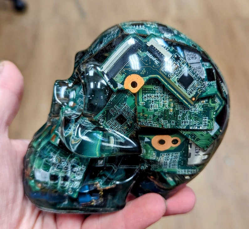 May include: A clear resin skull sculpture with embedded green circuit boards. The skull is detailed with intricate circuitry and has a glossy finish.