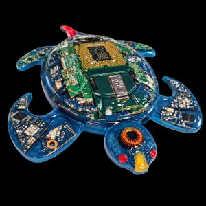 May include: A turtle-shaped sculpture crafted from blue resin, featuring embedded electronic components including a central CPU socket, green circuit boards, and a copper coil. The head includes two red circular accents and a yellow beak-like feature.