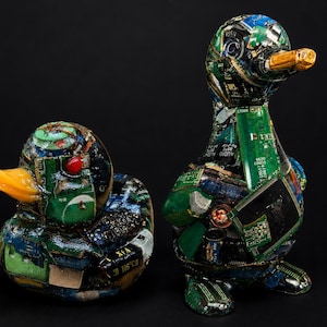 May include: Two duck sculptures constructed from recycled green and blue electronic circuit boards. The ducks feature glossy yellow beaks and intricate patterns of integrated chips, capacitors, and metallic components covering their entire surfaces.