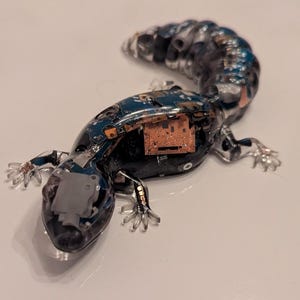 May include: A clear, resin sculpture of a lizard made from recycled circuit boards. The lizard is posed in a walking position with its tail curled up. The sculpture is a unique and interesting piece of art.