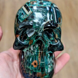 May include: A green and black skull sculpture made of resin with embedded circuit boards. The skull is detailed with a textured surface and a glossy finish.