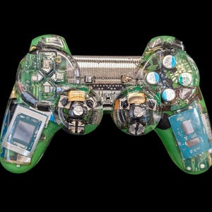 May include: A green and clear resin-encased video game controller, showcasing internal electronic components. The controller features visible circuit boards, chips, and buttons, set against a black background. The design highlights the intricate technology within.