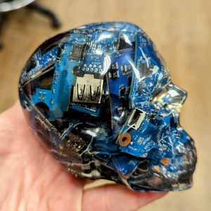 May include: A clear resin skull filled with blue and silver circuit boards. The skull is a decorative object and is made of epoxy resin.