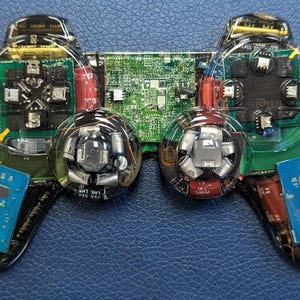 May include: A transparent video game controller with exposed internal components, including circuit boards, wires, and buttons. The controller has a clear plastic shell, revealing the intricate electronic parts. The color scheme includes green, blue, red, and silver elements.