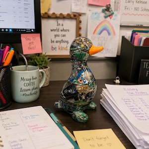 May include: A decorative duck figurine covered in a collage of recycled circuit board fragments with a bright orange beak, positioned on a desk among office supplies, a mug, a planner, and papers with handwritten text including Essay Due Friday.