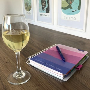 May include: A wine glass filled with white wine sits on a wooden surface, next to a spiral-bound planner and a pen. In the background, a bottle of wine and framed travel art prints are visible.