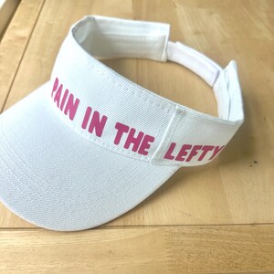 Pode incluir: Viseira branca com o texto "PAIN IN THE LEFTY" em letras rosa. A viseira tem uma aba curva e uma alça ajustável. A viseira é feita de tecido.