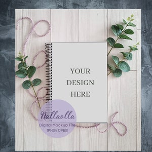 以下が含まれることがあります： 薄い木の背景に、ピンクのビーズと緑のユーカリの葉が飾られた、白いスパイラルバインディングのノート。ノートには「YOUR DESIGN HERE」という文字が書かれています。