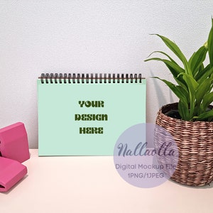 May include: A light green spiral bound notebook with the text "YOUR DESIGN HERE" in dark green. The notebook is on a white surface with a pink binder clip and a potted plant with green leaves in a brown wicker pot.