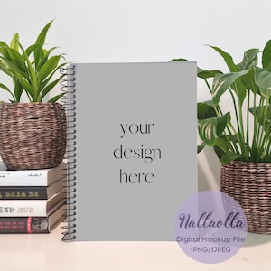 Può includere: Un mockup di un quaderno a spirale grigio con il testo "your design here" sulla copertina. Il quaderno è appoggiato su un tavolo con due piante in vaso, una su ogni lato. Le piante sono in cesti intrecciati marroni.