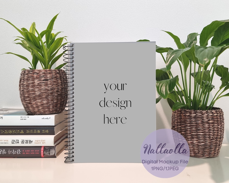 May include: A gray spiral bound notebook mockup with the text "your design here" on the cover. The notebook is sitting on a table with two potted plants, one on each side. The plants are in brown woven baskets.