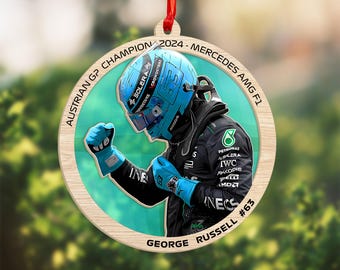 May include: Wooden Christmas ornament featuring George Russell, #63, in a racing suit and helmet. The ornament has a circular design with the text "AUSTRIAN GP CHAMPION 2024 - MERCEDES AMG F1" around the edge. A red ribbon is attached for hanging.