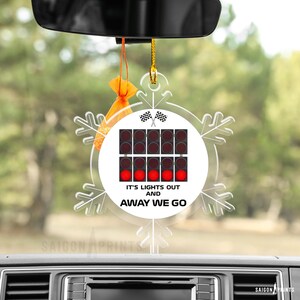May include: A clear, snowflake-shaped car ornament with the text "IT'S LIGHTS OUT AND AWAY WE GO." The ornament features a graphic of race starting lights and checkered flags. An orange air freshener hangs above.