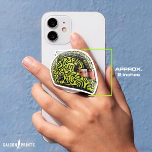 Op de afbeelding: Een telefoonhouder met een racehelm-ontwerp met neon groene en zwarte abstracte patronen. De houder is ongeveer 5 cm groot. De afbeelding bevat ook de tekst "SAIGON PRINTS".