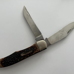 May include: A brown handled pocket knife with two blades, one open and one closed. The knife has a wood-like handle with a pattern.