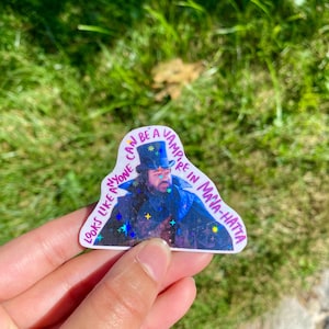 May include: A white sticker with a glittery background and a picture of a man in a black coat and top hat. The text on the sticker reads "Can anyone look like a vampire in Maya-Hatta"