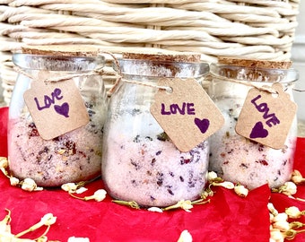 Peut inclure: Trois pots en verre remplis de sels de bain, avec des couvercles en liège et des étiquettes « Love » avec un cœur violet. Les pots sont sur du papier rouge, avec des fleurs séchées.