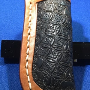 May include: A brown leather knife sheath with a white stitching detail and a black embossed pattern. The sheath is designed to hold a pocket knife.