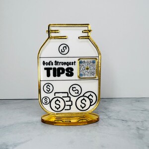 May include: A gold-colored, decorative jar with a white label that reads "God's Strongest TIPS". The jar has a QR code and dollar signs on the label. The jar is on a gold-colored stand.