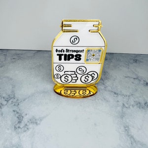 May include: A gold-colored, decorative jar with a white label that reads "God's Strongest TIPS". The jar has a QR code on the label and is surrounded by gold dollar signs.