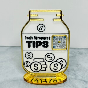 May include: Gold-colored metal piggy bank with a white label that reads "God's Strongest TIPS" and a dollar sign. The piggy bank has a QR code and several dollar signs on the front.