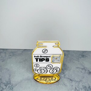 May include: A gold-colored acrylic tip jar with a white label that reads "God's Strongest TIPS". The jar is filled with gold coins and has a gold base.
