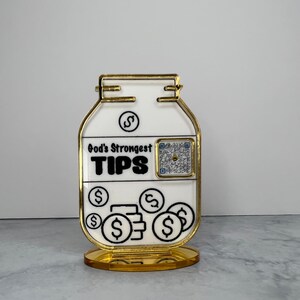 May include: A gold-colored glass jar with a white label that reads "God's Strongest TIPS". The jar has a gold-colored base and a QR code on the front. The jar is filled with gold-colored coins with dollar signs on them.