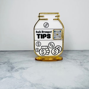 May include: A white ceramic jar with a gold rim and base. The jar has a black and white design with the text "God's Strongest TIPS" and a QR code. The jar is filled with gold dollar signs.