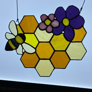 May include: A stained glass bee and honeycomb design with purple, pink, and yellow flowers. The honeycomb is made of yellow and orange glass.