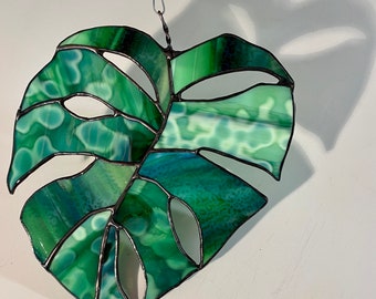 May include: A stained glass window hanging with a green monstera leaf design. The leaf is made of multiple pieces of glass with a dark green, light green, and blue color scheme.