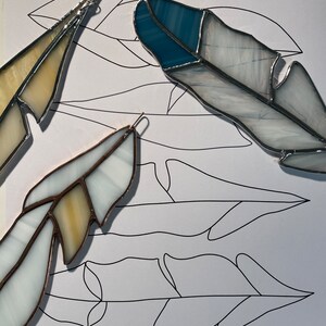 Pode incluir: Três penas de vitral em tons de branco, azul e amarelo, com bordas de metal prateado. As penas estão sobre uma superfície branca com contornos de penas. O texto na parte inferior diz "Fawn Beach Glass 2022 Feather Set."