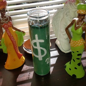 May include: A green candle with a white dollar sign and the number 20 on it. The candle is surrounded by decorative figurines of women in colorful dresses and a white angel statue.