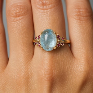 May include: A gold ring with a large, oval, pale blue gemstone at its centre. Small, round, red gemstones are set either side of the main stone. The ring is on a person's finger.