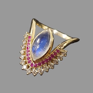 May include: A gold ring featuring a marquise-cut, blue gemstone at the center. The ring is accented with small, round, red and white gemstones, creating a decorative, symmetrical design. The ring has a unique, openwork design.