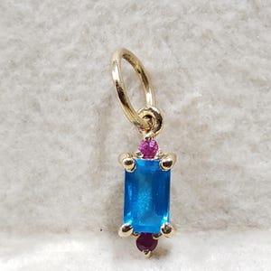 May include: A gold pendant charm with a blue rectangular gemstone surrounded by four smaller pink gemstones.