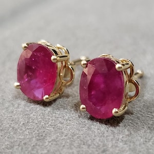 Op de afbeelding: Een paar gouden oorstekers, elk met een ovaal geslepen robijn. De robijnen hebben een diepe rode kleur en zijn gezet in een delicate gouden zetting. De oorbellen worden gepresenteerd op een grijze ondergrond.