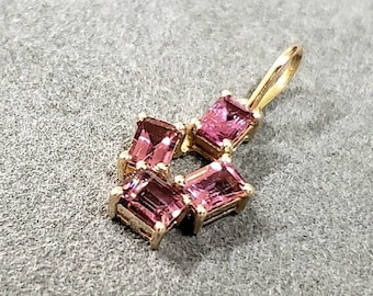 May include: A gold pendant with five pink square-shaped gemstones. The pendant is on a gray textured surface.