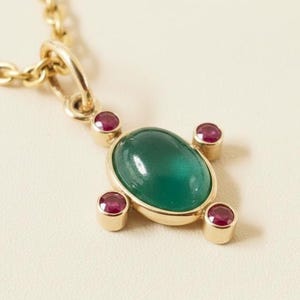 May include: A gold necklace featuring a pendant with a large, oval, green gemstone at the center, surrounded by four smaller, round, red gemstones. The pendant is suspended from a delicate gold chain, creating an elegant jewelry piece.