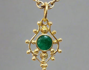 May include: A gold-coloured necklace with a pendant featuring a round, emerald-green gemstone. The pendant has an ornate design with small, gold-coloured spheres. The chain is also gold-coloured and has a delicate link pattern.